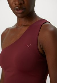 Detailopname van een persoon in een mouwloos bordeauxrood top met een Puma-logo op de borst, zichtbaar zijn de nek, schouder en lippen.