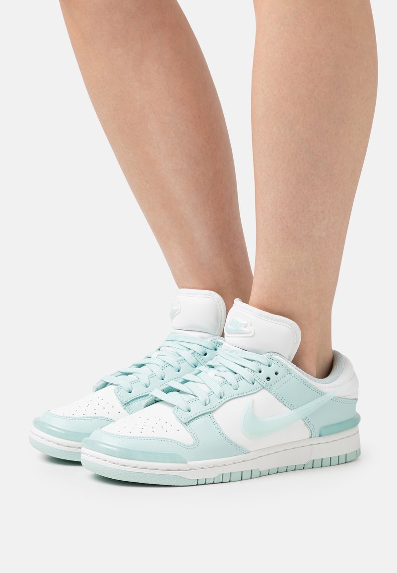 Personne portant des baskets Nike blanches et bleu clair avec des lacets, debout devant un fond blanc uni.