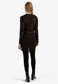 Donna che indossa una tunica nera trasparente a maniche lunghe con motivo floreale e cintura, leggings neri e stivaletti neri con tacco, in piedi di spalle.