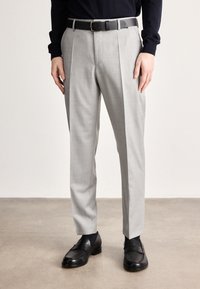 Calças cinza claro feitas à medida com textura suave, apresentando pregas na frente e um cinto preto. Combinadas com sapatos de couro escuro e polido.