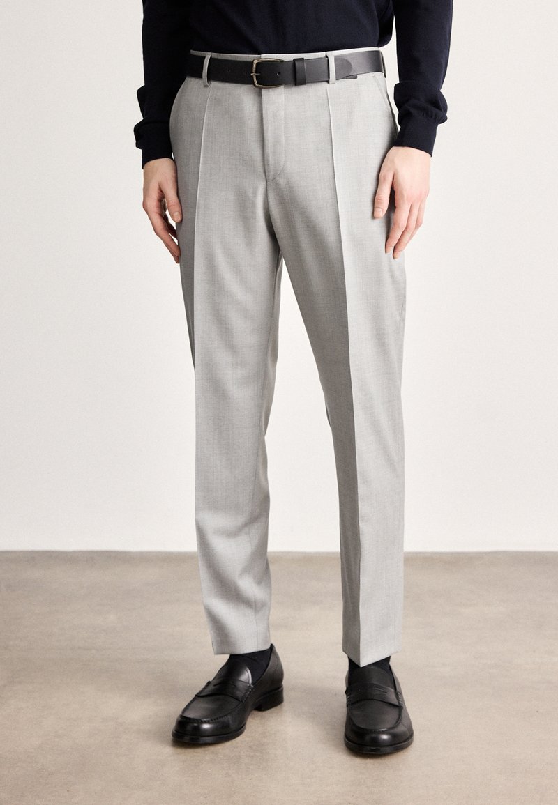 Calças cinza claro feitas à medida com textura suave, apresentando pregas na frente e um cinto preto. Combinadas com sapatos de couro escuro e polido.