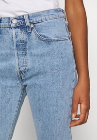 Primer plano de unos pantalones vaqueros de mezclilla azul claro y parte de una camisa blanca, mostrando el bolsillo delantero, la cintura, el botón y una mano derecha en reposo.