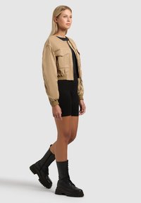 Femme debout portant une veste courte beige, un short noir ajusté et des bottes de combat noires à lacets sur un fond gris clair.