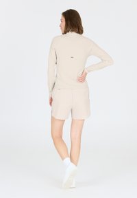 Beige Sportoberteil mit langen Ärmeln und einem hohen Kragen, kombiniert mit passenden Shorts. Dezente gerippte Textur mit kleinen goldenen Akzenten.