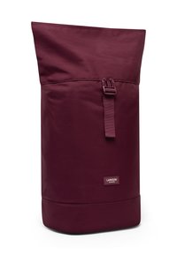 Zaino roll-top color marrone realizzato in tessuto resistente. Presenta una chiusura anteriore a fibbia e una piccola etichetta con il marchio vicino al fondo.