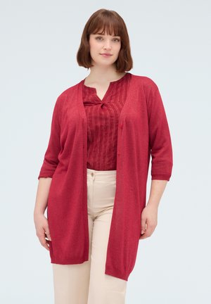 Frau mit kurzen braunen Haaren, die einen langen roten Cardigan, eine rote Bluse mit dezenten Streifen und hellbeige Hosen trägt, steht vor einem schlichten Hintergrund.