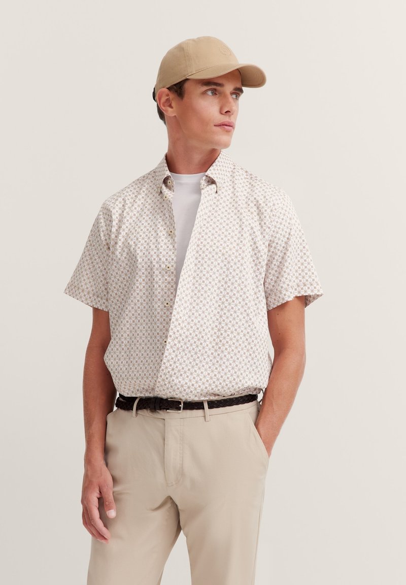 Kurzärmliges Hemd mit Knöpfen in Weißgrund und geometrischem braunem Muster. Kombiniert mit beigen Hosen und einer sandfarbenen Cap. Glatte Stofftextur.