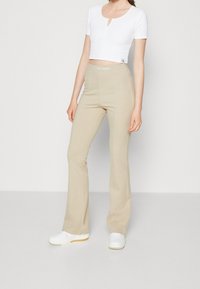 Žena v béžových kalhotách s rozšířenými nohavicemi od Calvin Klein Jeans, bílém zkráceném tričku s krátkým rukávem a bílých teniskách, stojící na neutrálním pozadí.