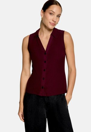 Femme portant un haut sans manches bordeaux foncé à boutons avec col et un pantalon noir, debout avec une main dans la poche.