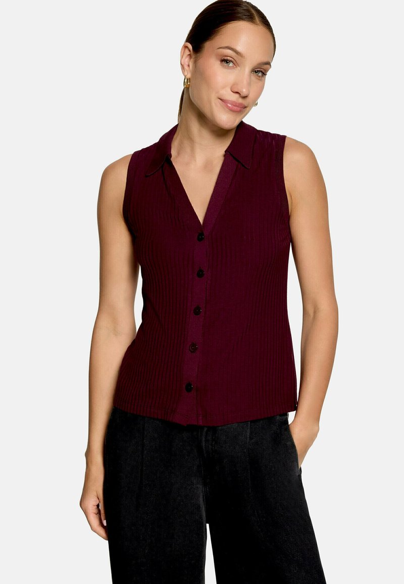 Femme portant un haut sans manches bordeaux foncé à boutons avec col et un pantalon noir, debout avec une main dans la poche.