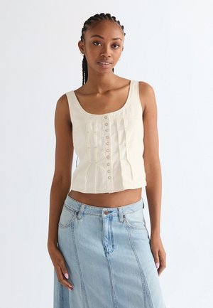 Frau mit Zöpfen, die ein ärmelloses, cremefarbenes, geknöpftes Oberteil und einen hellblauen Jeansrock trägt, stehend vor einem schlichten weißen Hintergrund.