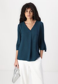 Teal, v-halsbluse med trekvart ermer, laget av teksturert stoff. Kombinert med hvite bukser og en sølvfarget clutch-veske.