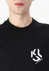 Black cotton t-shirt featuring a round neckline and white graphic logo on the upper left side. Smooth texture, casual fit.