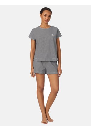 Jonge vrouw staat op blote voeten, draagt een zwart-wit gestreept topje met korte mouwen en bijpassende korte broek, neutrale gezichtsuitdrukking, effen witte achtergrond.