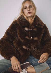 Hnedá, chlpatá bunda s veľkým golierom, zapínaním na šnúrky a mäkkou textúrou. Nosená cez rifle a hnedú obuv.