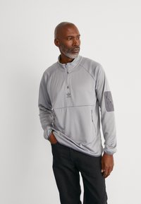Halv-zip, lys grå genser med strukturert stoff, adidas-logo på brystet, glidelåslomme på ermet, og kontrasterende grå stoffdetaljer.