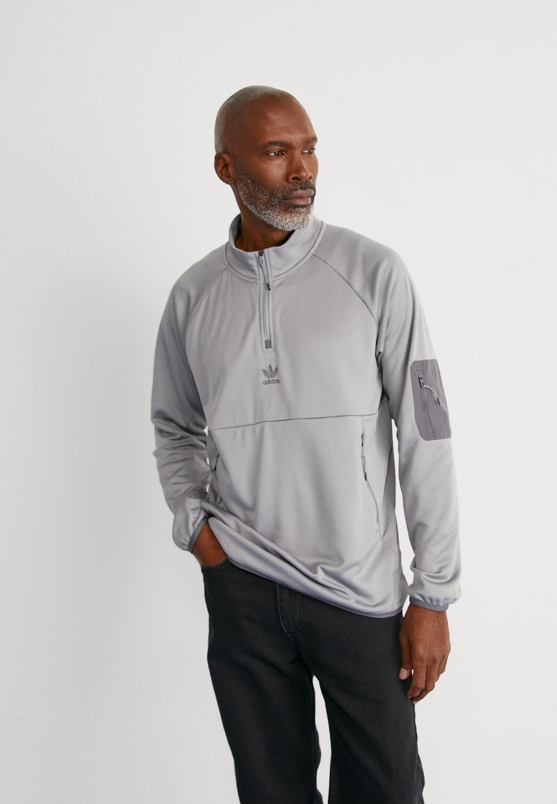 Halv-zip, lys grå genser med strukturert stoff, adidas-logo på brystet, glidelåslomme på ermet, og kontrasterende grå stoffdetaljer.