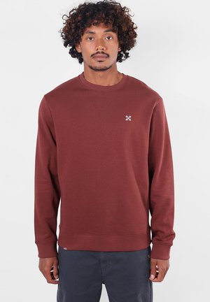 Homme aux cheveux bouclés portant un sweat-shirt uni bordeaux à manches longues et un pantalon foncé, debout devant un fond clair.