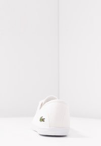 Baskets en toile blanches avec une finition texturée, présentant un logo crocodile vert Lacoste sur le talon et une semelle en caoutchouc blanche.