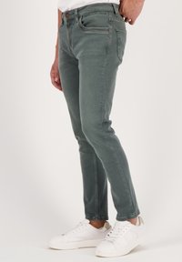 Slim-fit vihreät farkut, joissa on etutaskut ja vaaleanruskea nappi, yhdistettynä valkoisiin tennareihin neutraalilla taustalla.