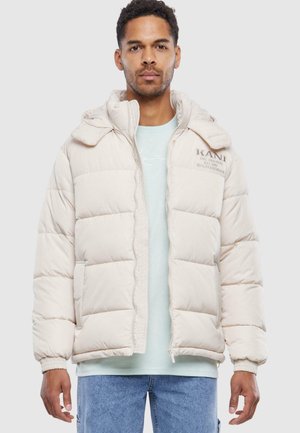 Chaqueta hinchable beige con diseño acolchado, capucha y cierre de cremallera. Presenta bordado en el pecho y dos bolsillos laterales. Textura suave.
