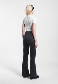 Valkoinen ribattu toppi lyhyillä hihoilla, yhdistettynä korkeavyötäröisiin, leveisiin mustiin farkkuihin. Malli seisoo selkä kameraan päin, näyttäen sauman yksityiskohtia.