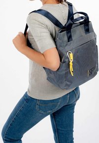 Marinblå strukturerad ryggsäck med dubbla handtag och justerbara remmar. Har en gul accent med en karbinhake. Bärs på ryggen över en grå topp.