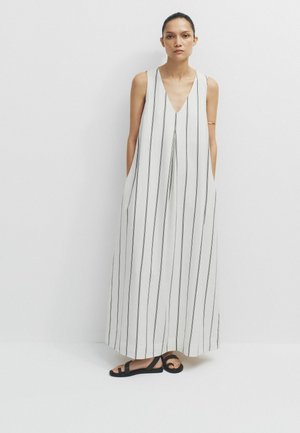 Woman wearing sleeveless white maxi dress with black vertical stripes and black flat sandals, standing against plain white background.