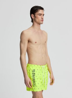Giovane uomo in piedi senza maglietta, che indossa pantaloncini da bagno giallo brillante con piccoli motivi e la scritta "TERRANOVA" sulla gamba sinistra.