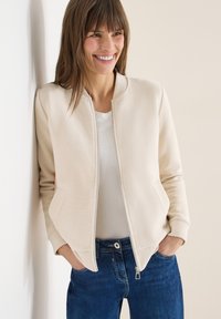 Femme souriante portant une veste bomber en côte crème sur une chemise blanche et un jean bleu, debout les mains dans les poches contre un mur uni.