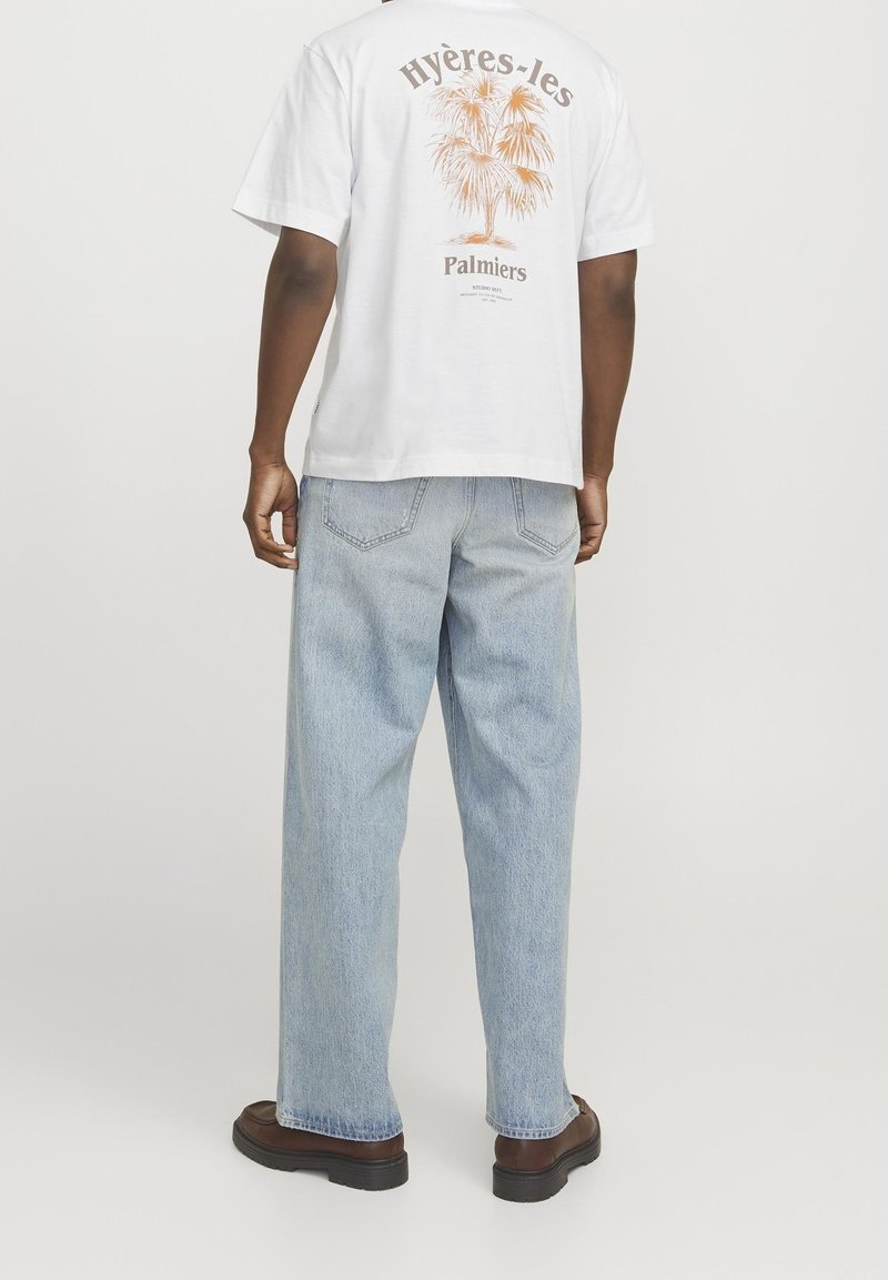 Person, die ein weißes T-Shirt mit einem orangefarbenen Palmbaum und dem Text "Hyères-les Palmiers" trägt, hellblaue Jeans und braune Schuhe, steht mit dem Rücken zur Kamera.