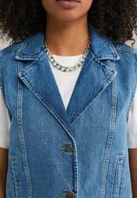 Denimväst i ljusblått med slagkrage, två framfickor och silverfärgade knappar. Bärs över en vit t-shirt med ett tjockt kedjehalsband.