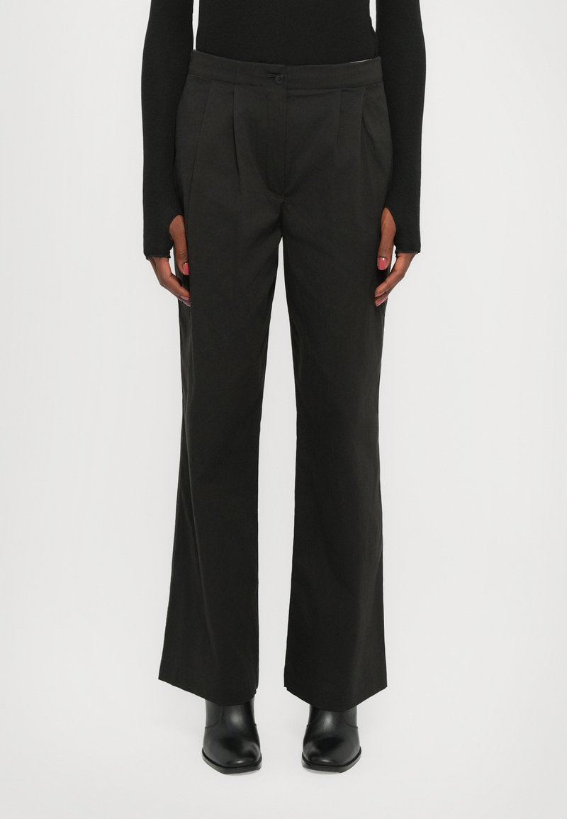 Pantalons noirs à jambes larges en tissu lisse, avec des plis avant et une fermeture à bouton à la taille, associés à des bottines noires.