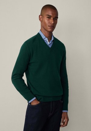 Grøn v-hals sweater lavet af strikstof, med lange ærmer og en afslappet pasform, lagdelt over en lyseblå skjorte med krave.