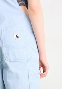 Hellblaue Latzhose aus strukturiertem Stoff mit einer Gesäßtasche und einem Logopatch. Das Design umfasst breite Schulterträger und eine lockere Passform.