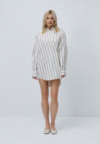 Witte jurk met verticale bruine strepen, lange mouwen, knoopsluiting aan de voorkant en zijsplitten. Zachte stof met een gladde textuur.