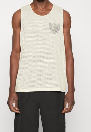 Débardeur en maille blanche sans manches avec un petit emblème sur le côté gauche de la poitrine, porté par une personne à la peau foncée et portant un short noir.