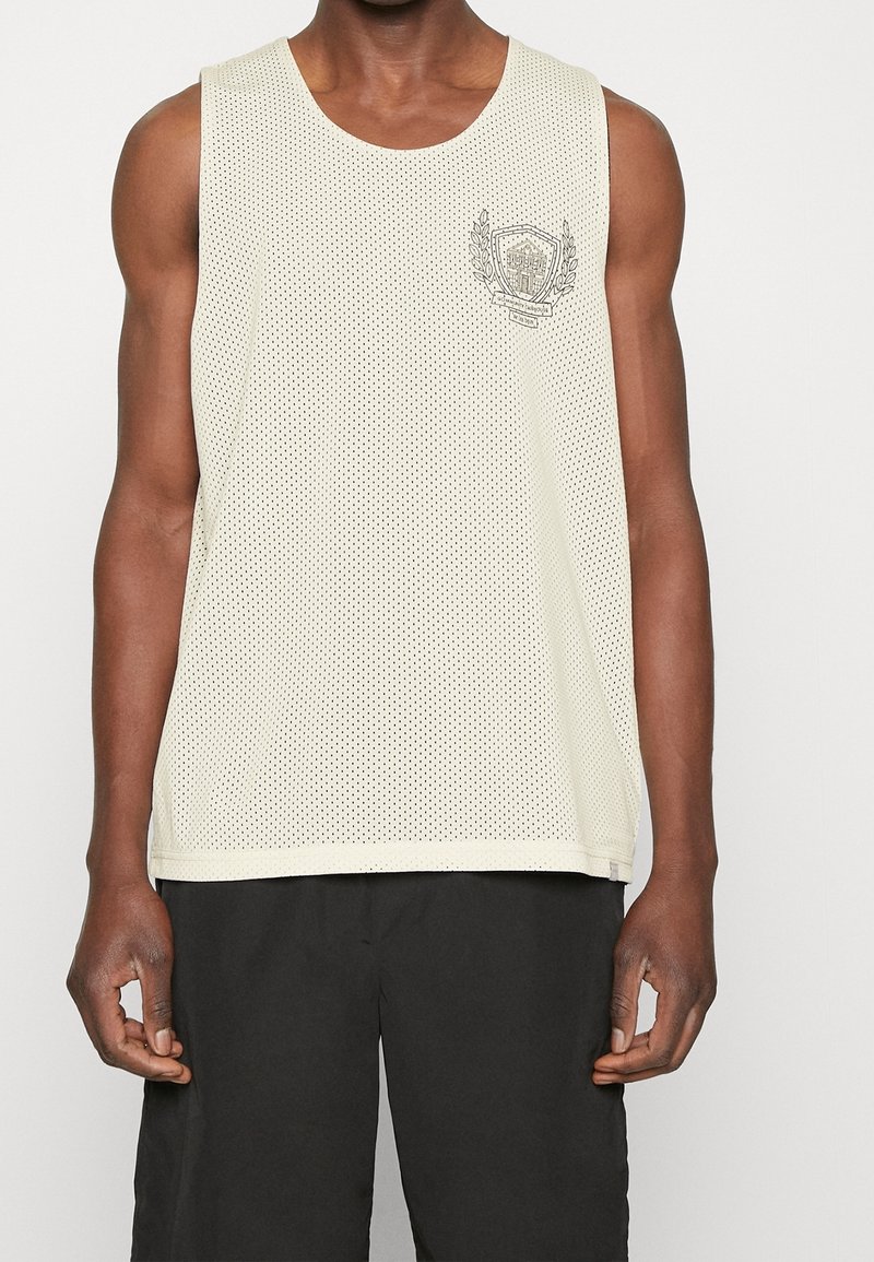Débardeur en maille blanche sans manches avec un petit emblème sur le côté gauche de la poitrine, porté par une personne à la peau foncée et portant un short noir.