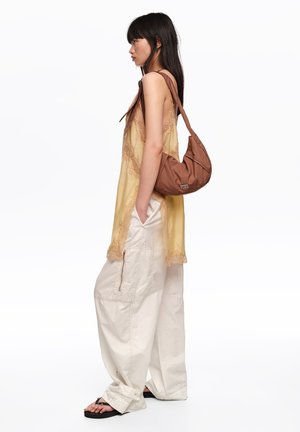 Joven mujer con pantalones cargo blancos y holgados y top amarillo de encaje, de pie de lado con bolso marrón al hombro y sandalias negras sobre fondo blanco.