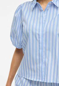 Camicia azzurra chiaro con strisce verticali bianche, maniche corte a sbuffo e colletto. Realizzata in un tessuto morbido.