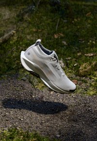 Chaussure de sport légère en blanc cassé avec une tige texturée, un design épuré et des accents subtils. Elle possède une semelle rembourrée et un laçage minimal.