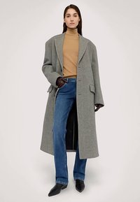 Abrigo gris de lana oversize con cuello solapa, llevado sobre un jersey de cuello alto beige. Combinado con vaqueros rectos azules y botas negras puntiagudas.
