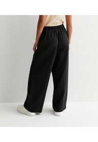 Svart vida byxor med elastisk midja, tillverkade av mjukt tyg, med en slät yta och avslappnad passform. Bärs med vita skor.