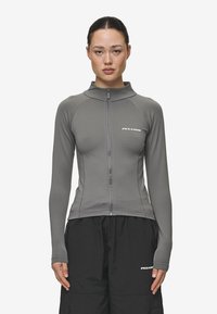 Pilkas zip-up džemperis, pagamintas iš lygaus audinio, su pamoksliniu apykakliu ir ilgomis rankovėmis, derinamas su juodomis laisvomis kelnėmis.