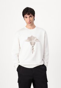 Sudadera blanca con un motivo gráfico beige que recuerda a una hoja estilizada. Está confeccionada con tejido suave, tiene un escote redondo y un corte relajado.
