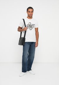 Mand iført hvid trykt t-shirt, blå jeans, hvide sneakers og bærende en sort skuldertaske, der står mod en ensfarvet hvid baggrund.