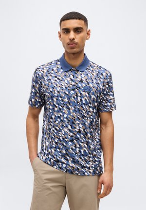 Homme portant un polo à motifs abstraits bleu, beige, noir et blanc avec un col bleu et un pantalon kaki, debout avec une main dans la poche.