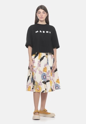 Donna in piedi di fronte indossando una T-shirt nera oversize con la scritta "MARNI", gonna midi floreale e scarpe platform color senape su sfondo bianco.