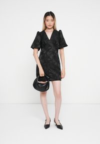 Čierne šaty s kvetinovým vzorom, s balónovými rukávmi, výstrihom do V a krátkym strihom. Modelka drží čiernu koženú kabelku a má na sebe čierne špicaté topánky.