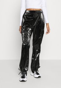 Black, shiny vinyl pants with a loose fit, featuring side pockets and drawstring hems. Paired with black and white sneakers.